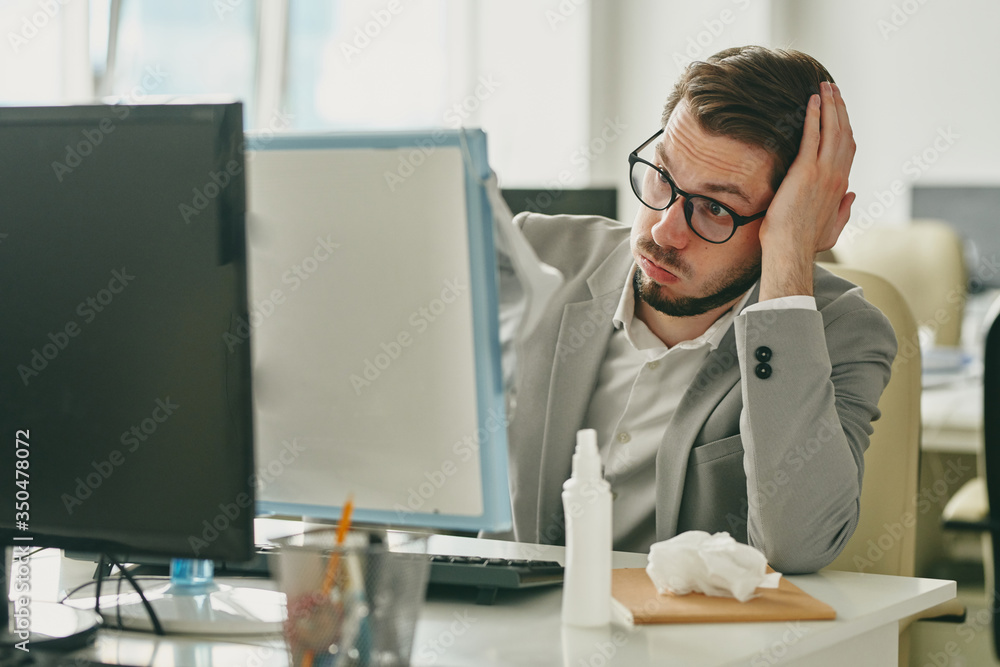Poster irritated young businessman in eyeglasses sitting at desk and looking at computer screen while havin