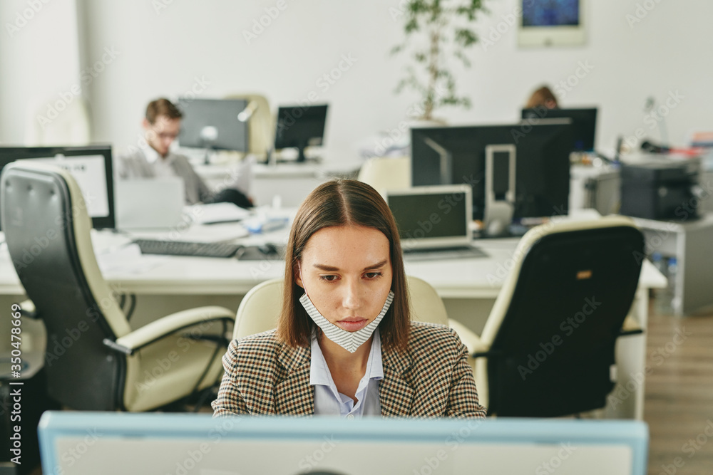 Poster Attractive young woman with mask on chin using modern computer while working in half-empty office during quarantine