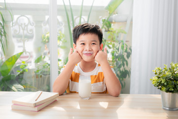 Adorable happy young Asian boy having breakfast and drinking milk at home in the morning; looking at camera - obrazy, fototapety, plakaty