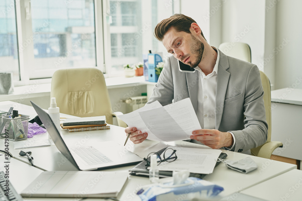 Poster focused young bearded businessman sitting at desk with laptop and examining contract while solving w