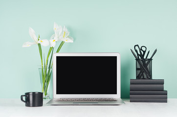 Simple minimalistic office interior with blank laptop monitor, black stationery, books, coffee cup, white iris flowers in green mint menthe interior on white wood table.