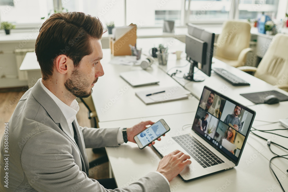 Poster Young bearded businessman working alone in office while connecting colleagues via video chat on laptop during coronavirus epidemic