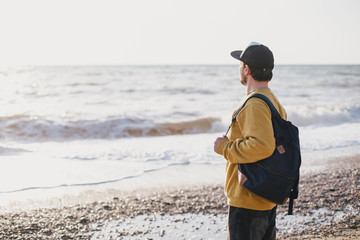 Young man traveler with beard wearing backpack exploring empty beach near ocean or sea.
