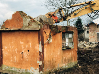 Excavator destroying brick house on land in countryside. Bulldozer clearing land from old bricks and concrete from walls with dirt and trash. Backhoe machinery ruining house