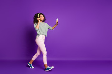 Fototapeta na wymiar Full size photo of positive afro american girl break pause go walk listen music choose song cellphone have ear phones wear striped shirt pink trousers sneakers isolated purple color background