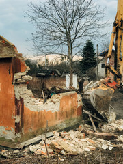 Fototapeta premium Excavator destroying brick house on land in countryside. Bulldozer clearing land from old bricks and concrete from walls with dirt and trash. Backhoe machinery ruining house