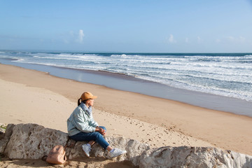 Young girl with a backpack sitting on the sandy beach watching the ocean waves of Costa Da Caparica, Portugal.