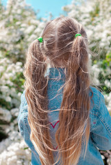Beautiful little girl with long blond hair with two ponytails in blue denim jacket on blooming nature background. The child standing back view alone in the flowering bushes park. Street photography.