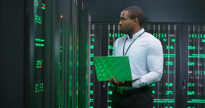 Male system administrator of big data center typing on laptop computer while working in server room. Programming digital operation. Man engineer working online in database center. Telecommunication.