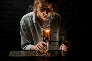 Beauty woman bartender holds tweezers with smoking stick near wineglass and looks at it