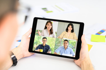 Back view of businessman has a meeting with Asain colleagues about plans in a video conference via tablet. New normal for working from home business team using online video call during quarantine