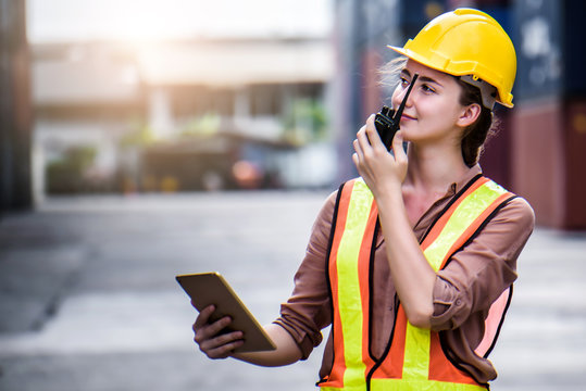 Young Confident Caucasian Woman Engineer Using Radio Communication And Wearing Yellow Safety Helmet And Check For Control Loading Containers Box From Cargo Freight Ship For Import And Export