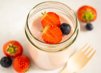 Dessert strawberry panna cotta in a glass jar decorated with cut strawberries and blueberries. On a light gray table with strawberries and a wooden dessert fork
