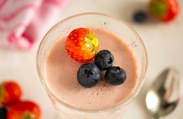 Dessert strawberry panna cotta in a glass wine glass is decorated with a whole strawberry and blueberry. On a light gray table with strawberries, a spoon and a napkin