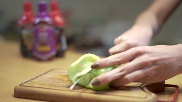Woman cuts green paprika on a wooden mat. Big knife and brown nails.