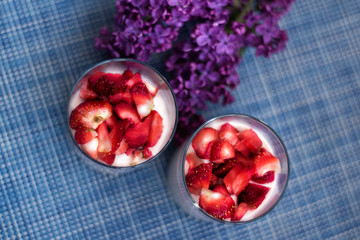 Layered strawberry homemade italian creamy dessert panna cotta with fresh berries in glass on blue background with branch of purple lilac. Organic sweet. Top view of strawberry panna-cotta