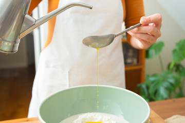 Cook in apron adding olive oil with spoon to wheat flour in ceramic bowl. Dough preparation for tasty pastry and bread. Studio shot. Side view. Homemade bakery and cooking at home concept