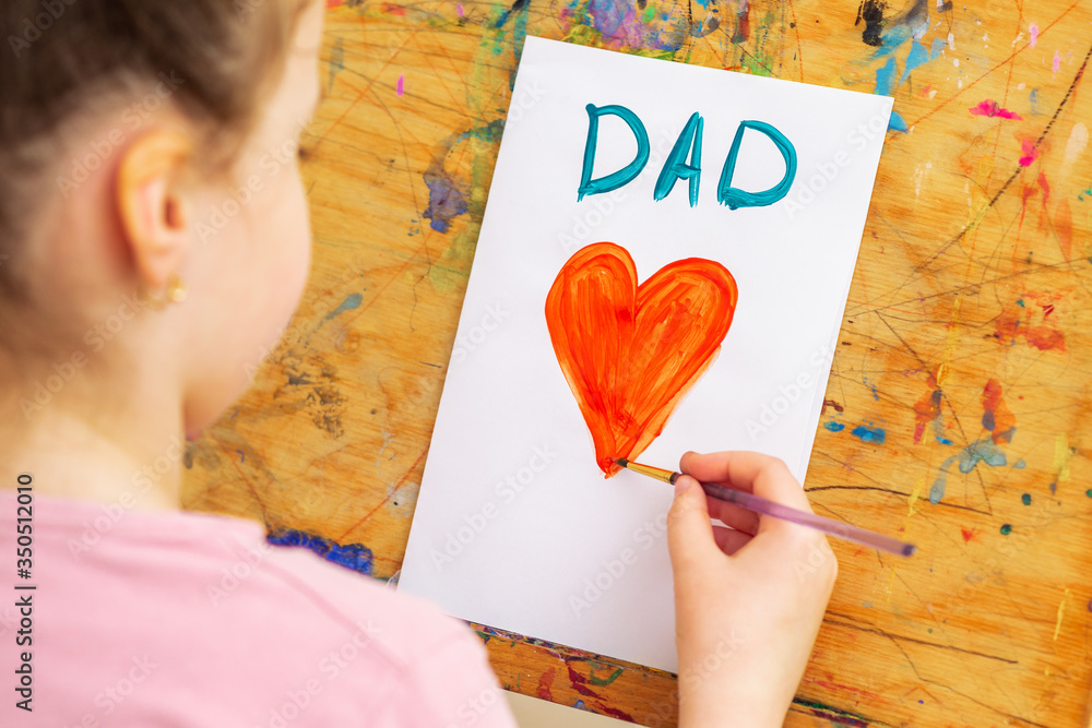 Wall mural Back view of littel girl is drawing red heart with word Dad greeting card by watercolors on white paper on an easel. Father's Day concept.