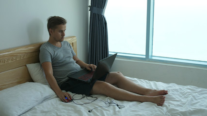 Young man sitting on bed using his laptop in the bedroom at home