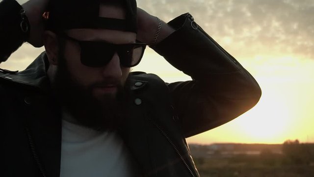 Bearded stylish biker motorcyclist puts on black cap at sunset in slow motion. Trendy fancy man in glasses and leather jacket. Brutal fashionable person with earring. Chic modish Life style. Biker.