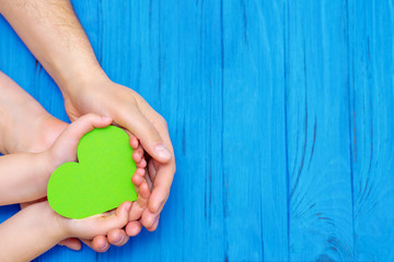 Top view of male and child hands holding paper green heart on wooden blue background. Concept of...