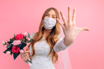 A bride in a wedding dress and a medical protective mask on her face on a pink background. Wedding, quarantine, coronavirus