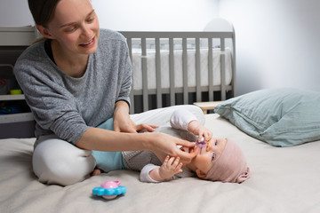 Obraz na płótnie Canvas Positive young mom giving soother to baby daughter, soothing child in bedroom. Mother and little child staying at home. Child care or quarantine concept