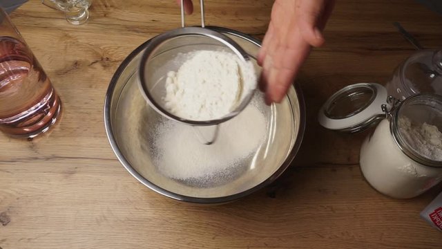 Chef is preparing flour for making a dough by sifting it through a metal sieve to a cooking bowl
