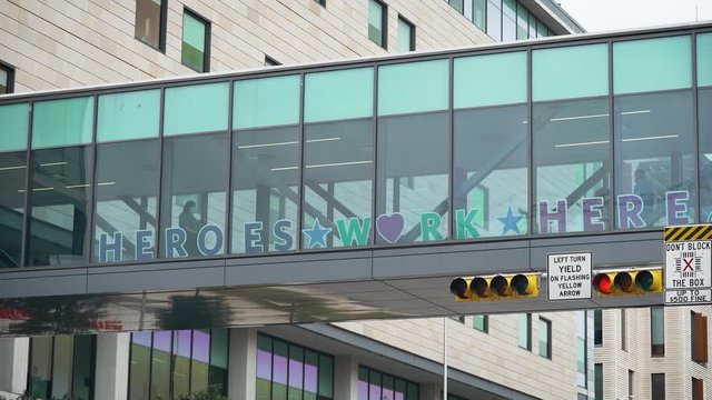 Heroes Work Here Sign On Glass Hospital Bridge With Nurses And Doctors Wearing Face Masks Walking By