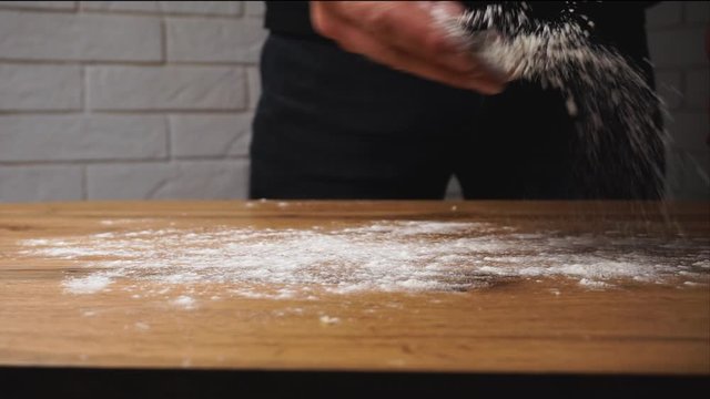 Chef pours a white dry flour across the table and than put a piece of fresh dough for kneading with strong hands