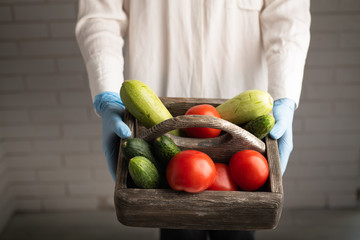 Woman in medical gloves holding a donation food box with tomatoes, zucchini and cucumbers