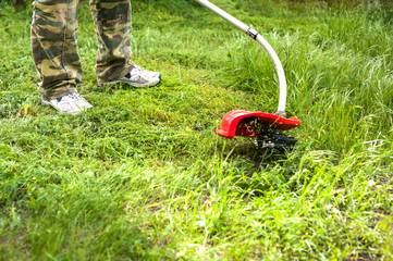 A man in a protective overalls mows the grass with a trimmer on a sunny day.
