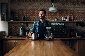 Smiling Man prepares coffee in the style of "pour over" in the barista school. Against the background of coffee equipment and a brick wall