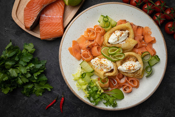 Pancakes with salmon, cheese, cucumbers and tomatoes on a dark background
