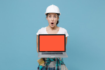 Shocked woman in protective helmet hardhat hold laptop pc computer with blank empty screen isolated on blue wall background. Instruments accessories for renovation apartment room. Repair home concept.