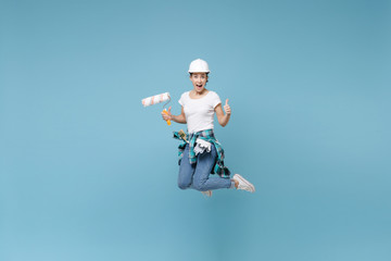 Obraz premium Excited young woman in protective helmet hardhat jump with paint roller isolated on blue wall background. Instruments accessories for renovation apartment room. Repair home concept. Showing thumb up.