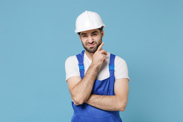 Confused young man in coveralls protective helmet hardhat isolated on pastel blue background. Instruments accessories for renovation apartment room. Repair home concept. Put hand prop up on chin.