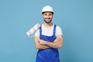 Smiling man in coveralls protective helmet hardhat hold paint roller isolated on blue background....