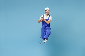 Fototapeta na wymiar Cheerful man in coveralls protective helmet hardhat hold electric drill isolated on blue background studio portrait. Instruments accessories renovation apartment room. Repair home concept. Jumping.