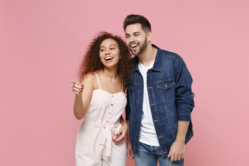 Cheerful young couple two friends european guy african american girl in casual clothes posing isolated on pastel pink background studio portrait. People lifestyle concept. Pointing index finger aside.