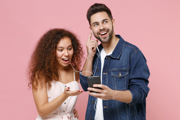 Excited young couple friends european guy african american girl in casual clothes posing isolated on pastel pink background. People lifestyle concept. Listen music with earphones using mobile phone.