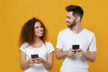 Smiling young couple friends european guy african american girl in white t-shirts isolated on yellow background. People lifestyle concept. Using mobile phone typing sms message, looking at each other.