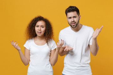 Perplexed puzzled young couple two friends european guy african american girl in white t-shirts posing isolated on yellow background. People lifestyle concept. Mock up copy space. Spreading hands.