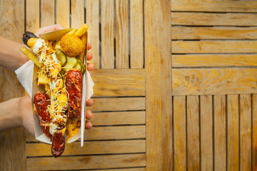 Obraz na płótnie Canvas Hands with a fork and street fast food on the background of a wooden table. Sausage, cheese, potatoes, sauce in a kraft plate. Street European fast food. Vertical.