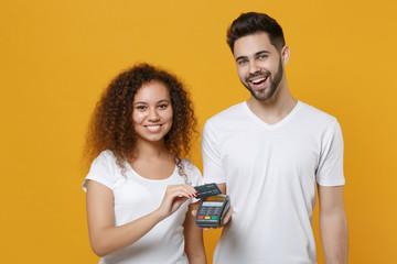 Smiling couple friends european guy african american girl in white t-shirts isolated on yellow background. People lifestyle concept. Hold bank payment terminal to process acquire credit card payments.
