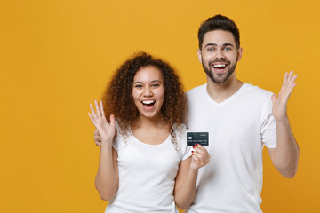 Excited young couple two friends european guy african american girl in white t-shirts posing isolated on yellow background in studio. People lifestyle concept. Hold credit bank card, spreading hands.