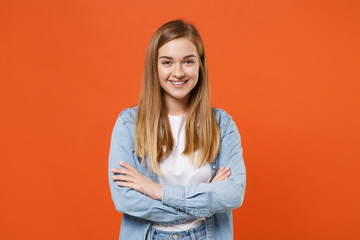 Smiling young woman girl in casual denim clothes posing isolated on bright orange wall background studio portrait. People sincere emotions lifestyle concept. Mock up copy space. Holding hands crossed.