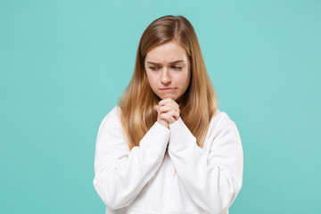 Pensive young woman girl in casual white hoodie posing isolated on blue turquoise background studio portrait. People emotions lifestyle concept. Mock up copy space. Holding hands folded in prayer.