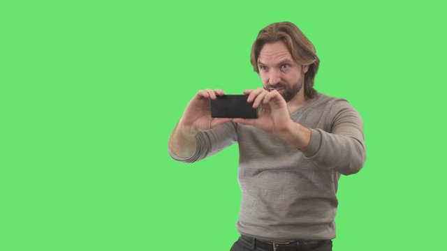 Brunette young man taking photos on smartphone. Portrait of Caucasian photographer using device on green background. Lifestyle, hobby, creativity. Chromakey, green screen.
