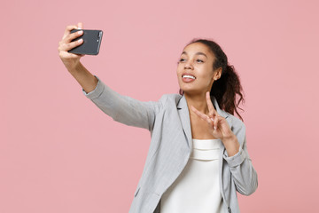 Smiling african american business woman in grey suit, white shirt isolated on pink wall background. Achievement career wealth business concept. Doing selfie shot on mobile phone, showing victory sign.